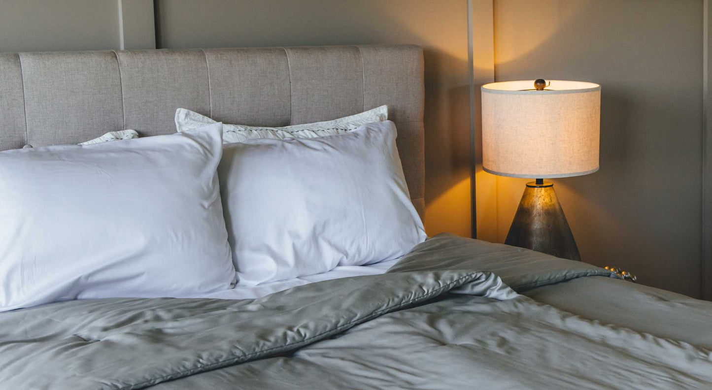 Neatly made bed with gray split comforter and white pillows, next to a lamp on a nightstand.